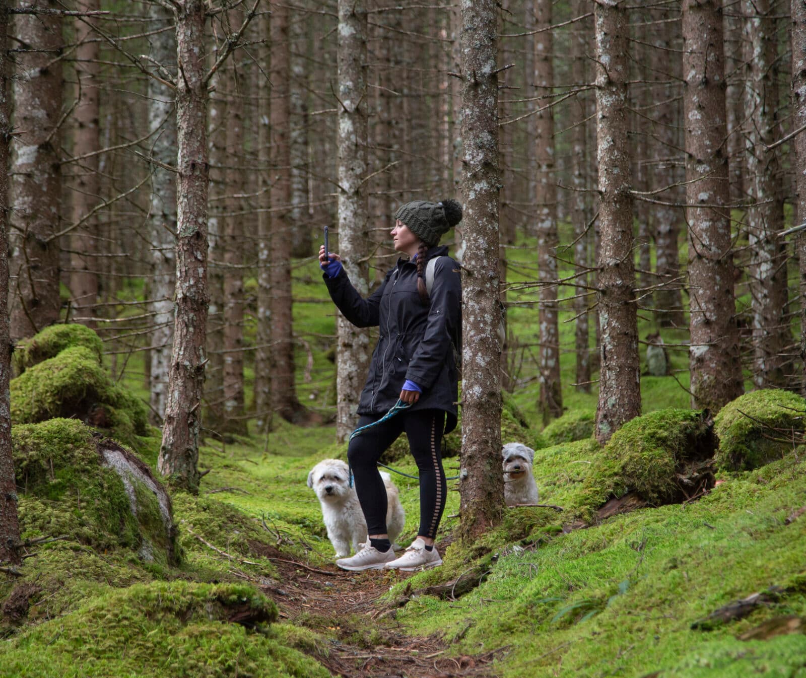 Kvinna står med hundar och mobiltelefon i skogen. Bilden har använts för digitala skogshjältar.