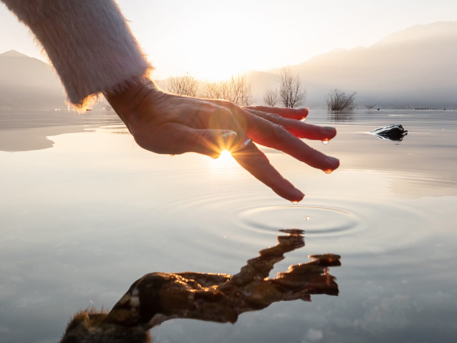 Detail of hand touching and caressing water surface of beautiful lake at sunset, mountain view. ...