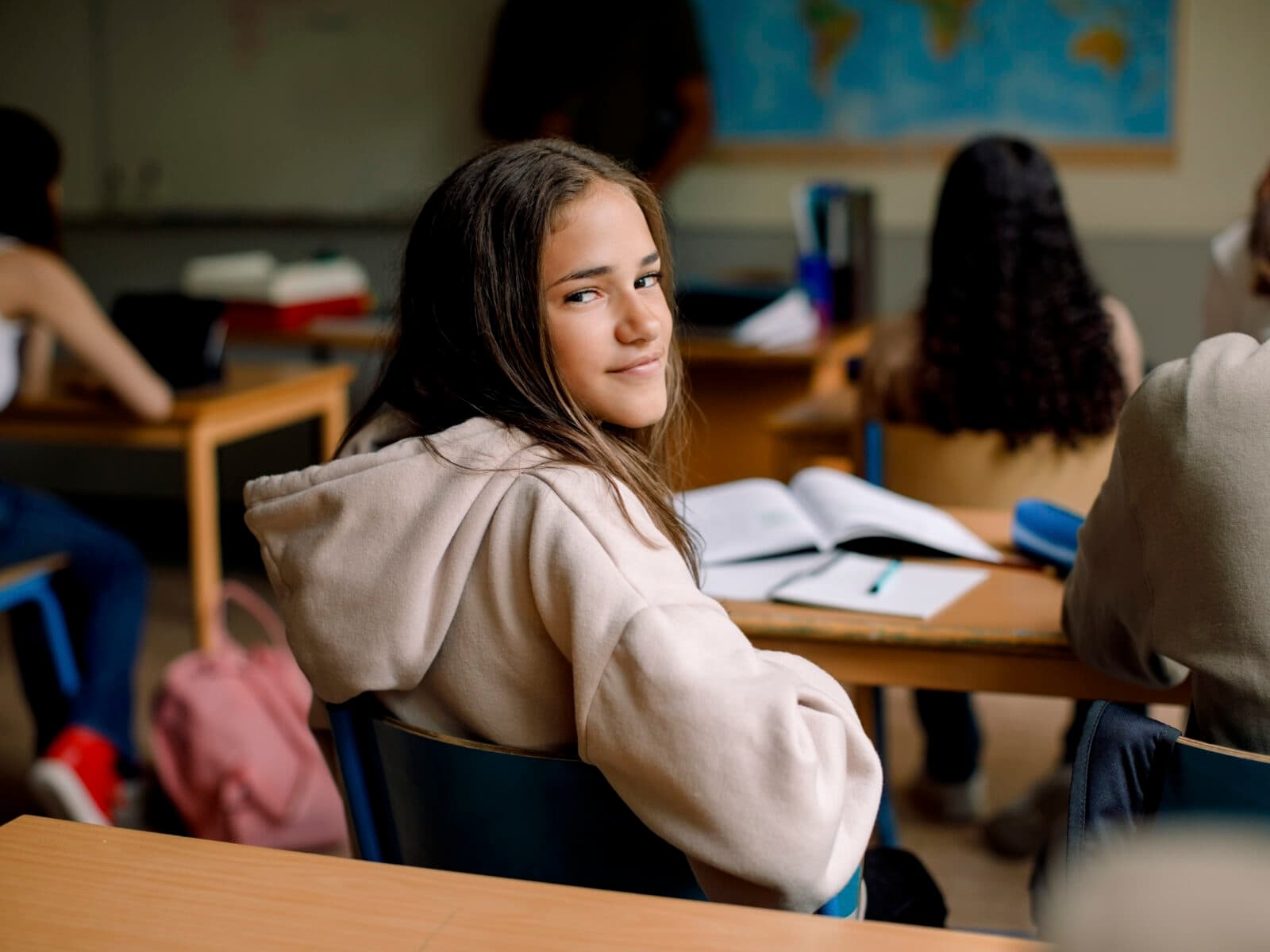 Bilden visar en klassrumsmiljö där en ung flicka sitter vid en skolbänk och tittar tillbaka mot kameran med ett halvt leende. Hon bär en beige huvtröja och hennes hår är löst. I bakgrunden kan man se andra elever som är fokuserade på sina skolarbeten och en världskarta som är upphängd på väggen, vilket antyder att det kan vara en geografilektion. Det finns böcker och skrivmaterial på borden, och atmosfären verkar lugn och studieinriktad.