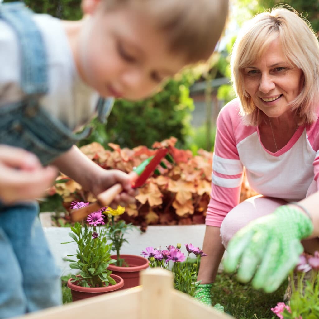 Bilden visar en kvinna och ett barn som odlar tillsammans utan gifter i en trädgård. Kvinnan, som har blont hår och bär rosa trädgårdshandskar, ler och sträcker sig mot en kruka med blommor. Hon ser ut att vara i 50-årsåldern och har på sig en randig tröja med rosa och vita ränder samt rosa byxor. Barnet, som är suddigt i förgrunden och verkar vara i förskoleåldern, bär hängselbyxor och håller i en liten spade eller trädgårdsredskap. De verkar vara i färd med att plantera eller sköta om växter i krukor, och det är en solig dag med grönska i bakgrunden. Atmosfären är avslappnad och ser ut att vara en trevlig stund i trädgården.