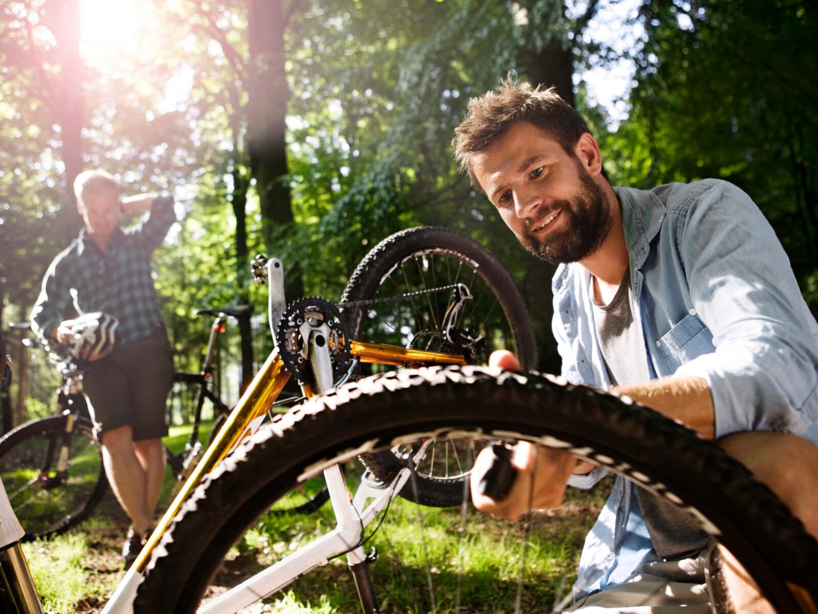 Använd i samband med Fröpengar. Bilden visar två män i en skogsmiljö under en solig dag. Mannen i förgrunden ler mot kameran och verkar vara i färd med att reparera eller justera ett cykeldäck på en mountainbike som ligger upp och ner. Mannen i bakgrunden står med en cykel bredvid sig och tittar på den andra mannen, möjligen väntar han på att reparationen ska bli klar. Området är grönt och lummigt, vilket tyder på att det är sommar eller vår. Ljuset som sipprar genom träden ger en varm och behaglig atmosfär.